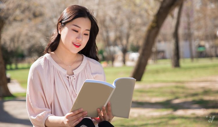 Pensive smiling college girl studying textbook in park. Young Asian woman reading book outdoors. Love reading concept